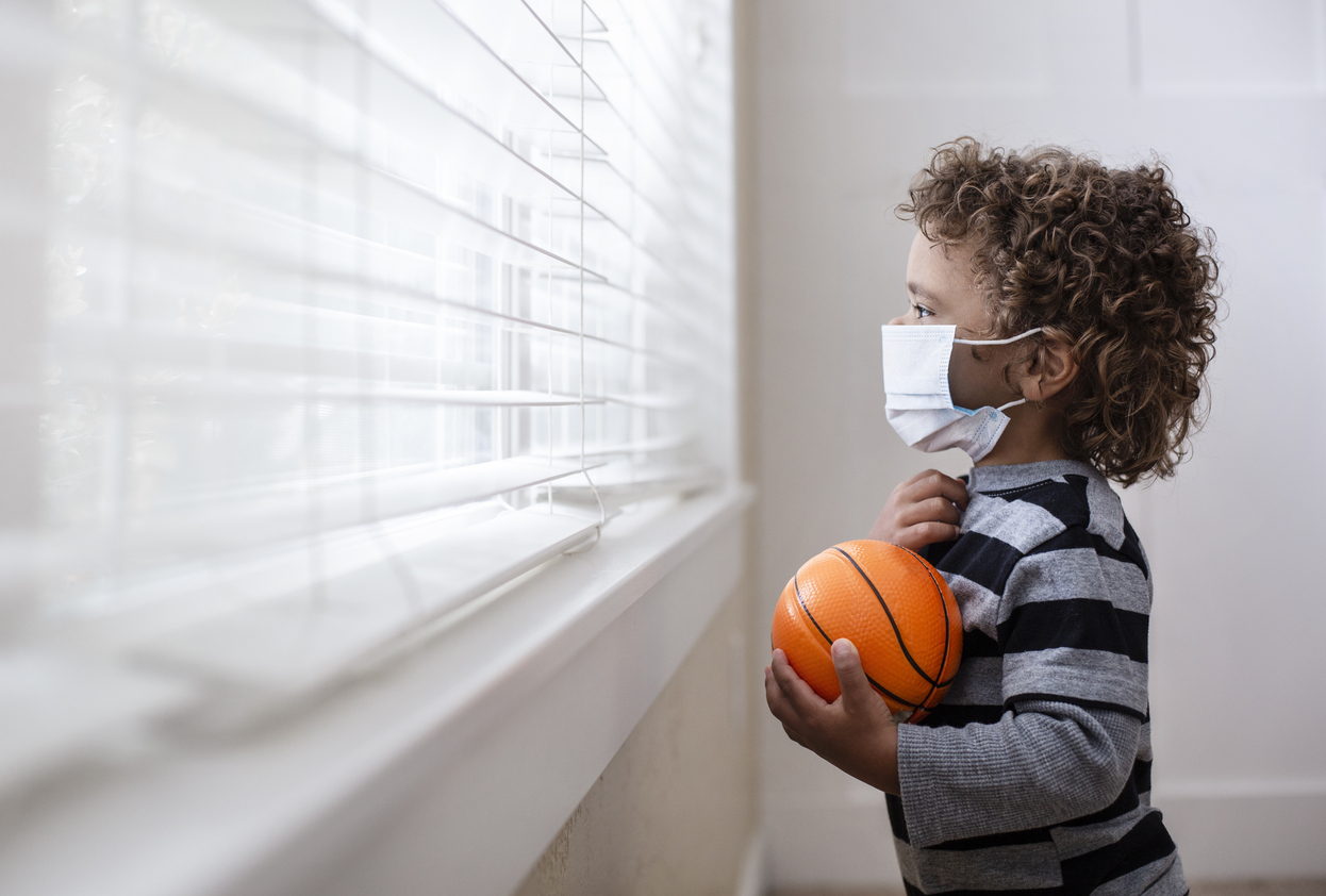 A young boy looking out the window wearing a protective facemark while seeking protection from COVID-19, or the novel coronavirus, by sheltering in place in his home.